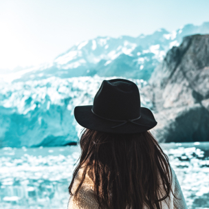 a person with long brown hair wearing a black fedora and white fur coat, looking at a glacier landscape